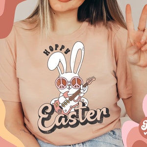 May include: A pink t-shirt with a graphic of a white bunny wearing pink sunglasses and playing a guitar. The bunny is holding a peace sign and the text "Hoppy Easter" is written in a retro font.