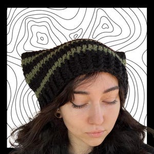 May include: A black and olive green crocheted beanie with cat ears. The hat features horizontal stripes and is worn by a person with long, dark hair. The background is a white and black abstract design.