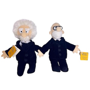 May include: Two plush dolls from the Brainy Beanies collection. The left doll features white hair, a mustache, and a black suit. The right doll has a bald head, a white beard, black glasses, and a black suit. Each doll has a yellow tag attached.