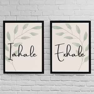 May include: Two framed prints with black frames. Each print displays a cursive word: "Inhale" and "Exhale". A botanical design in soft green and beige tones complements the text. The prints are designed for wall decor.
