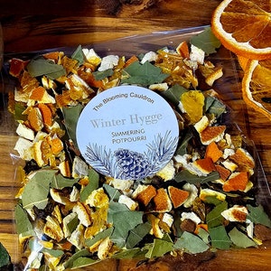 May include: A clear plastic bag filled with dried orange peel, bay leaves, and other spices. The bag has a round label that says "The Blooming Cauldron Winter Hygge Simmering Potpourri".