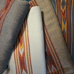 Puede incluir: Cuatro mantas tejidas con coloridos patrones geométricos. Las mantas están dobladas y apiladas una encima de la otra. La manta superior es azul, la siguiente es marrón, la siguiente es blanca y la manta inferior es gris.