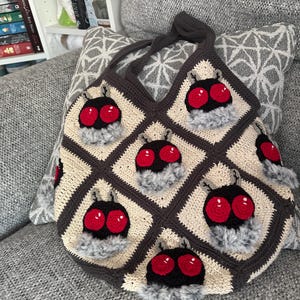 K&ouml;nnte beinhalten: Handgefertigte H&auml;keltasche mit einem sich wiederholenden Muster stilisierter Insektenk&ouml;pfe mit roten Augen und flauschigen grauen Akzenten. Die Tasche ist aus cremefarbenem, braunem und schwarzem Garn gefertigt und hat einen passenden braunen Griff.