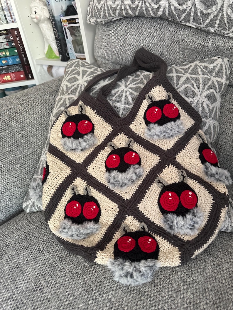 K&ouml;nnte beinhalten: Handgefertigte H&auml;keltasche mit einem sich wiederholenden Muster stilisierter Insektenk&ouml;pfe mit roten Augen und flauschigen grauen Akzenten. Die Tasche ist aus cremefarbenem, braunem und schwarzem Garn gefertigt und hat einen passenden braunen Griff.