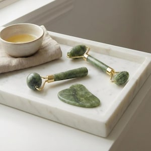 Op de afbeelding: Een spa-afbeelding met een witte marmeren tray met een kleine kom, een gevouwen beige handdoek en groene jade gezichtsrollers en een gua sha-tool. De rollers hebben goudkleurige metalen accenten.
