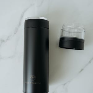 May include: Black stainless steel travel mug with a silver lid and a logo that says "Rich & Pour Co.". The mug is next to a clear plastic lid with a black base.
