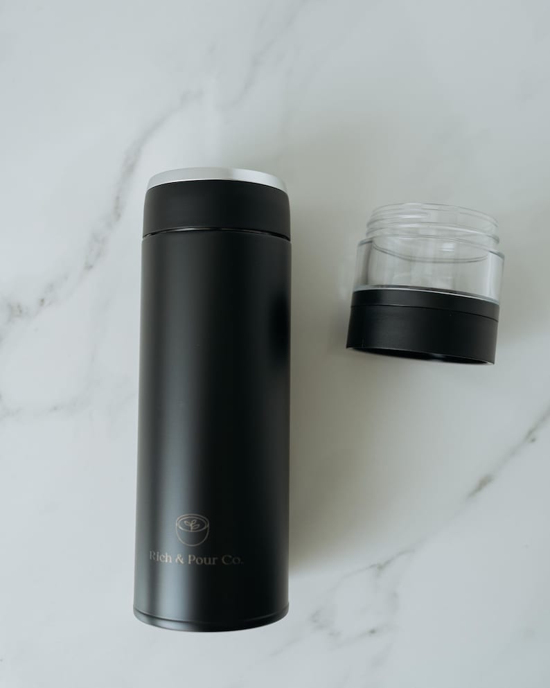 May include: Black stainless steel travel mug with a silver lid and a logo that says "Rich & Pour Co.". The mug is next to a clear plastic lid with a black base.