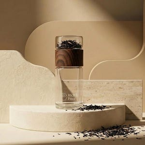 May include: A clear glass tea infuser with a dark wood band and lid, filled with loose tea leaves. The words "Rich & Pour" are printed on the glass. The infuser sits on a white platform, with scattered tea leaves around it.