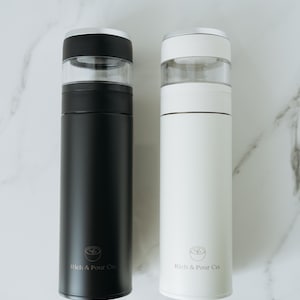 May include: Two stainless steel thermal coffee tumblers with clear lids. The tumbler on the left is black and the tumbler on the right is white. Both tumblers have the logo "Rich & Pour Co." printed on the side.