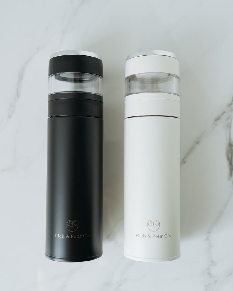 May include: Two stainless steel thermal coffee tumblers with clear lids. The tumbler on the left is black and the tumbler on the right is white. Both tumblers have the logo "Rich & Pour Co." printed on the side.