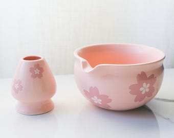 May include: A pink ceramic matcha bowl and whisk holder set. The bowl has a spout and is decorated with floral designs. The whisk holder is a small, rounded shape, also adorned with floral designs. Both items are on a white surface.