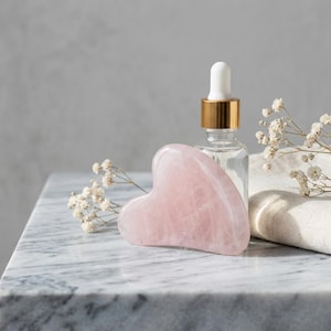 Op de afbeelding: Een roze rozenkwarts gua sha-tool, een heldere glazen fles met een gouden dop en een gevouwen witte doek zijn gerangschikt op een marmeren oppervlak. Delicate witte bloemen zijn rond de objecten geplaatst. De scène wordt verlicht door zacht, natuurlijk licht.