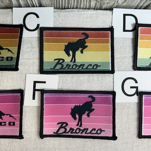 May include: Six embroidered patches with a retro color scheme. Three patches feature a silhouette of a Bronco truck driving over a mountain range. The other three patches feature a silhouette of a bucking bronco. All patches have the word "Bronco" written in a stylized font.