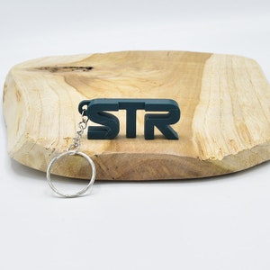 Puede incluir: Un llavero verde oscuro con las letras "STR" en estilo 3D. El llavero está unido a un anillo y una cadena de metal plateado.