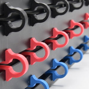 Puede incluir: Primer plano de una pared negra con filas de ganchos de plástico en negro, rojo y azul. Los ganchos tienen forma de U con un pequeño labio en la parte superior.