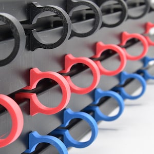 以下が含まれることがあります： 黒いプラスチック製のラックのクローズアップ。3列のカラフルなケーブルクリップが付いています。一番上の列には黒いクリップ、真ん中の列には赤いクリップ、一番下の列には青いクリップが付いています。