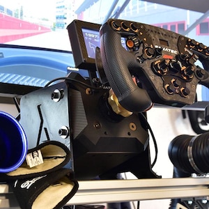 May include: A black racing steering wheel with multiple buttons and knobs mounted on a base, featuring a small digital display screen. Two black and blue air ducts are positioned on either side of the base, with a small control panel nearby.