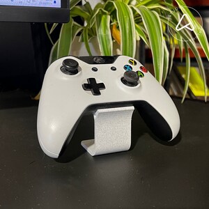 Op de afbeelding: Witte Xbox-controller met zwarte accenten op een witte standaard. De controller heeft een zwart D-pad, zwarte knoppen en zwarte triggers.