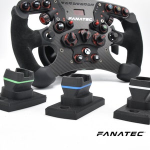 May include: A black racing steering wheel with red and silver accents, the word "FANATEC" printed on the front, and three black stands with green, blue, and white accents.