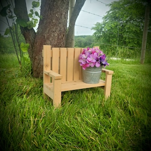 May include: A small wooden bench with a metal bucket filled with purple and pink flowers sitting on top of it. The bench is in a grassy area with a tree in the background.