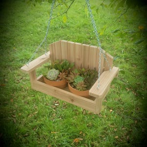 May include: A wooden hanging planter shaped like a small bench, suspended by chains. Two terracotta pots filled with green succulents sit inside. The planter is made of light-colored wood and hangs against a backdrop of green grass and foliage.