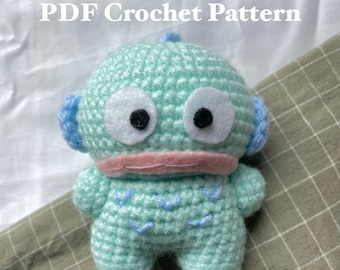 May include: A crocheted amigurumi toy in the shape of a light blue robot with large white eyes and a pink mouth. The toy is sitting on a green and white checkered fabric. The text "PDF Crochet Pattern" is at the top of the image and "@knotmichelle" is at the bottom of the image.
