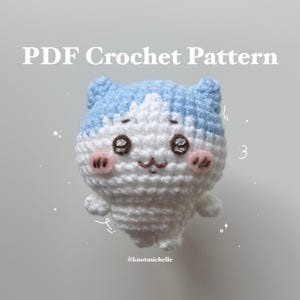 Puede incluir: Un juguete de gato tejido a ganchillo con un esquema de color azul y blanco. El gato tiene ojos marrones, mejillas rosadas y una pequeña sonrisa. El texto "PDF Crochet Pattern" está en la parte superior y "@knotmichelle" en la inferior.