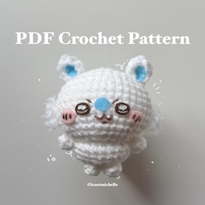 Könnte beinhalten: Ein weißer gehäkelter Bär mit blauen Akzenten an Ohren und Nase sowie rosa Wangen. Der Bär hat gestickte Augen und einen kleinen Mund. Der Text "PDF Crochet Pattern" steht oben und "@knotmichelle" unten.