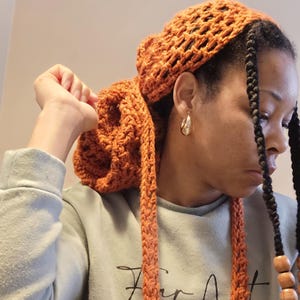 May include: A woman with long black braids is wearing a crocheted orange head scarf. The scarf is tied in a knot at the back of her head and the ends are hanging down her back.