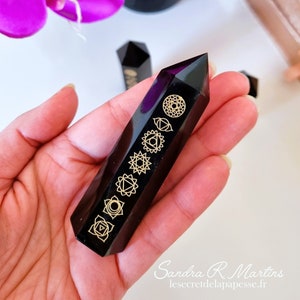 Peut inclure: Pointe de cristal d'obsidienne noire avec des symboles de chakras dorés gravés sur les côtés. Le cristal est tenu dans une main.