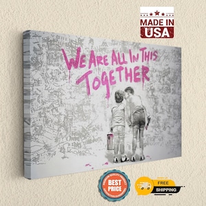 May include: A rectangular canvas wall art piece featuring a black and white illustration of two children standing before a wall covered in graffiti. The text WE ARE ALL IN THIS TOGETHER is printed in bright pink spray paint style across the top.