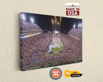 Può includere: Una stampa su tela rettangolare che mostra una vista aerea notturna di uno stadio di football gremito con fuochi d'artificio che esplodono sopra il campo. Il campo mostra il logo LSU viola e oro. Un badge MADE IN USA è visibile nell'angolo.