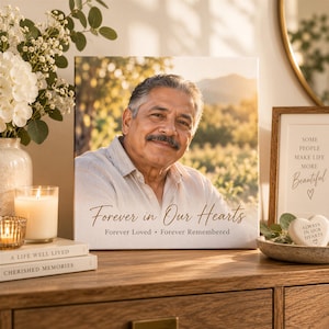 a wooden dresser with a framed portrait of an older man, a candle, a book, and a small bowl of flowers. The portrait has the text "Forever in Our Hearts Forever Loved Forever Remembered" written on it.