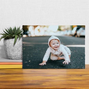A framed photograph of a smiling baby is displayed on a wooden surface, with a potted plant and a stack of books to the left.