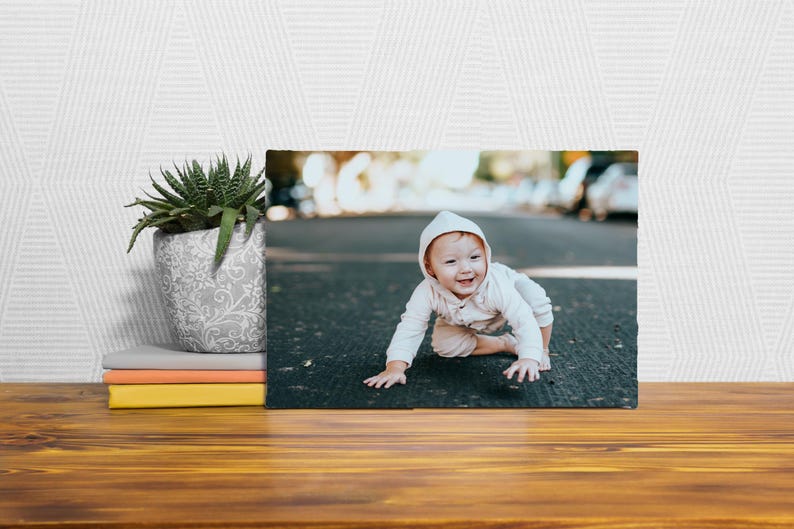 A framed photograph of a smiling baby is displayed on a wooden surface, with a potted plant and a stack of books to the left.