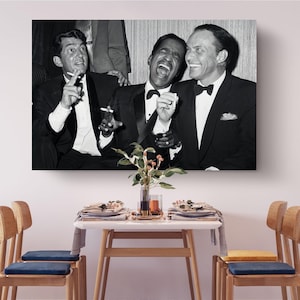 Könnte beinhalten: Ein schwarz-weiß Wandbild mit drei Männern in formellen Smokings, die lachen und Zigaretten halten. Das Bild hängt über einem Esstisch, der mit Tellern, Servietten und einer zentralen Vase mit Grünpflanzen gedeckt ist, flankiert von vier Holzstühlen.