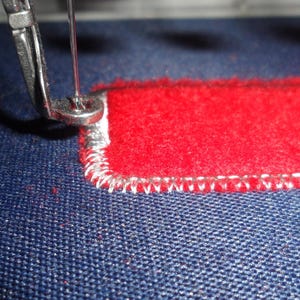 May include: Close-up of a sewing machine needle stitching a red rectangular patch onto blue fabric. The patch has a white stitched border. The needle is silver and the thread is white.