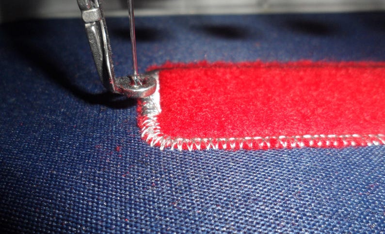 May include: Close-up of a sewing machine needle stitching a red rectangular patch onto blue fabric. The patch has a white stitched border. The needle is silver and the thread is white.