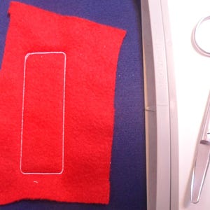 May include: A red felt rectangle with a white stitched outline, resting on a blue surface. A pair of silver scissors is positioned to the right. The image suggests a craft or sewing project.