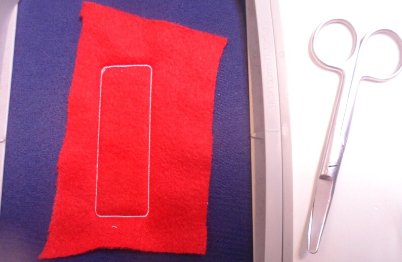 May include: A red felt rectangle with a white stitched outline, resting on a blue surface. A pair of silver scissors is positioned to the right. The image suggests a craft or sewing project.