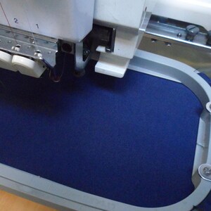 May include: An industrial embroidery machine with a navy blue fabric secured in a metal hoop. The machine's needle bar is visible, with multiple needles and thread spools. The machine is set up for detailed textile work.