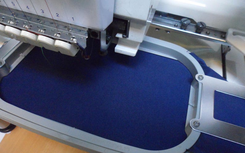 May include: An industrial embroidery machine with a navy blue fabric secured in a metal hoop. The machine's needle bar is visible, with multiple needles and thread spools. The machine is set up for detailed textile work.