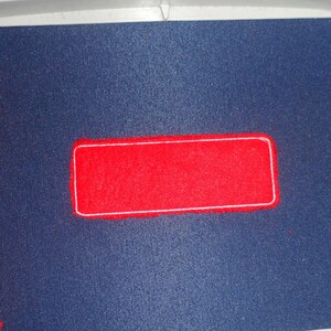 May include: A close-up of an embroidery project in progress. A red rectangular patch with a white border is centered on a dark blue fabric. Scissors are visible on the right side, and red fabric scraps are on the left.