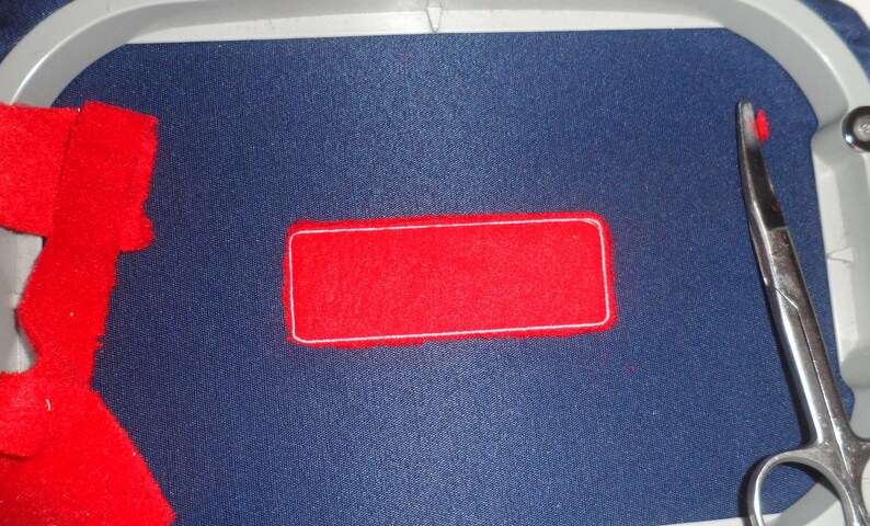 May include: A close-up of an embroidery project in progress. A red rectangular patch with a white border is centered on a dark blue fabric. Scissors are visible on the right side, and red fabric scraps are on the left.