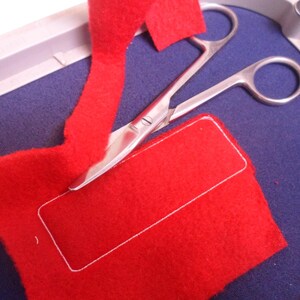 May include: A close-up shot shows a red felt fabric with a white stitched rectangle, and a pair of silver scissors resting on a blue surface. A strip of red felt is being cut. The scene suggests a crafting or sewing project.