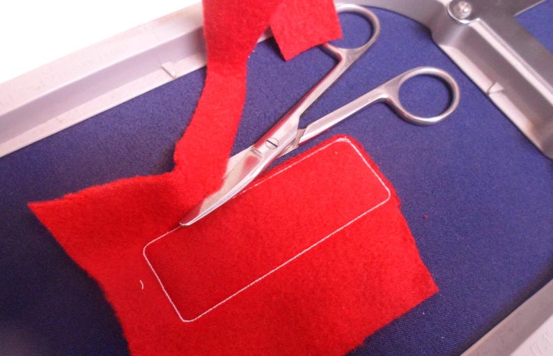 May include: A close-up shot shows a red felt fabric with a white stitched rectangle, and a pair of silver scissors resting on a blue surface. A strip of red felt is being cut. The scene suggests a crafting or sewing project.