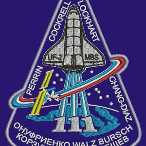 Könnte beinhalten: Ein dreieckiger Patch mit einem Raumschiff, das ins All startet. Der Patch ist weiß mit blauen und roten Akzenten. Der Text auf dem Patch lautet "PERRIN, COCKRELL, LOCKHART, UF-2, MBS, CHANG-DIAZ, 111, ОНУФРИЕНКО WALZ BURSCH, КОРЗУН WHITSON TРЕЩЕВ".