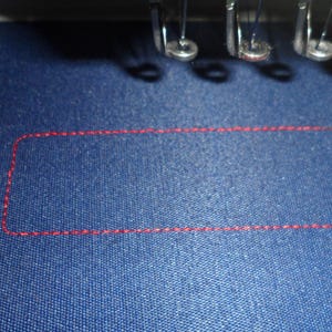 May include: Close-up of a blue fabric with a red stitched rectangular outline. The stitching is visible, and the fabric has a textured appearance. The image also shows parts of an embroidery machine.