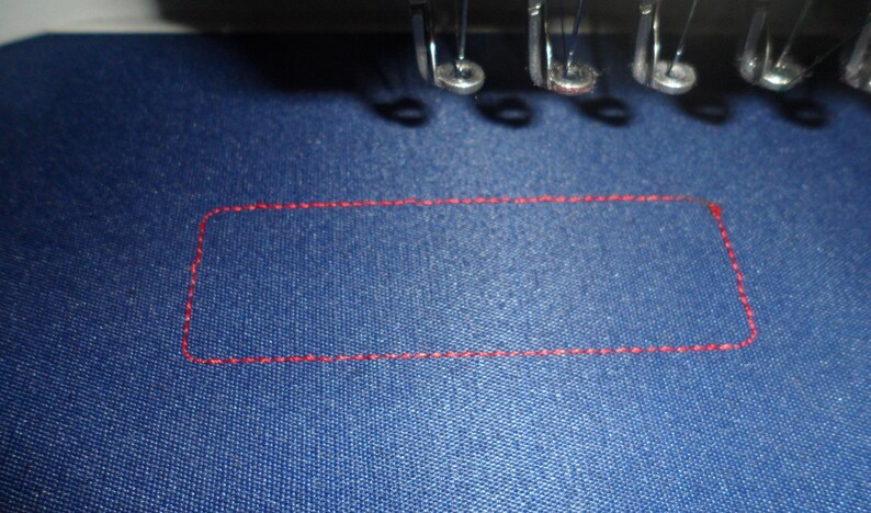 May include: Close-up of a blue fabric with a red stitched rectangular outline. The stitching is visible, and the fabric has a textured appearance. The image also shows parts of an embroidery machine.