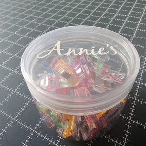 May include: A clear plastic container filled with colorful sewing clips. The container has a lid with the word "Annie's" printed on it. The clips are various colors, including pink, orange, green, and blue. The container is on a black cutting mat.